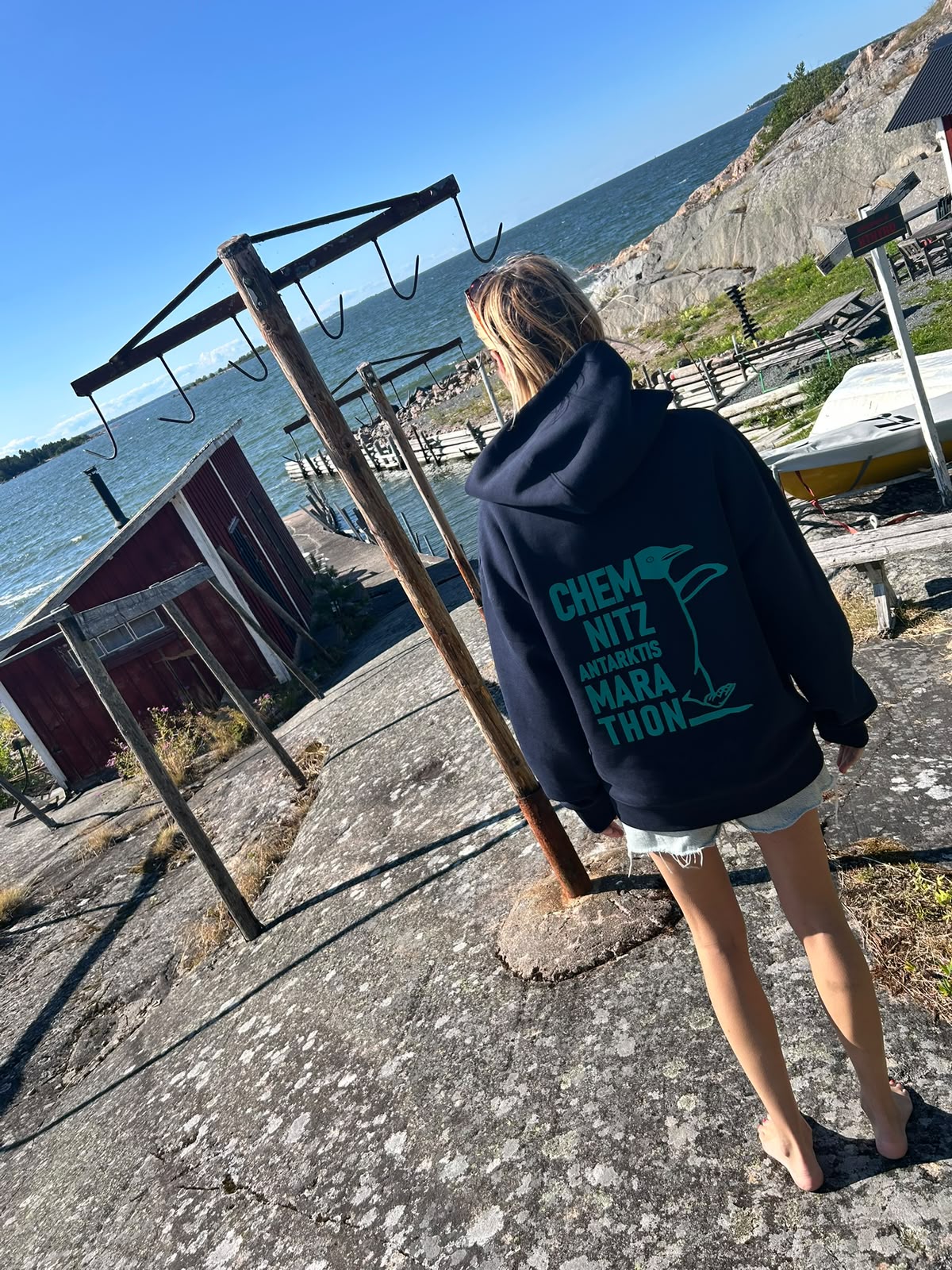 Person von hinten auf Felsen am Wasser, trägt den dunkelblauen Hoodie mit Rückendruck